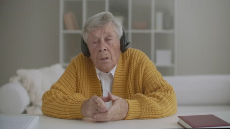 Großmutter-80-Jahre-Alt,-Ich-Schaue-In-Die-Kamera-Und-Rede,-Während-Ich-Mit-Kopfhörern-Sitze.-Fernkommunikation.-Fernanruf-Per-Videolink